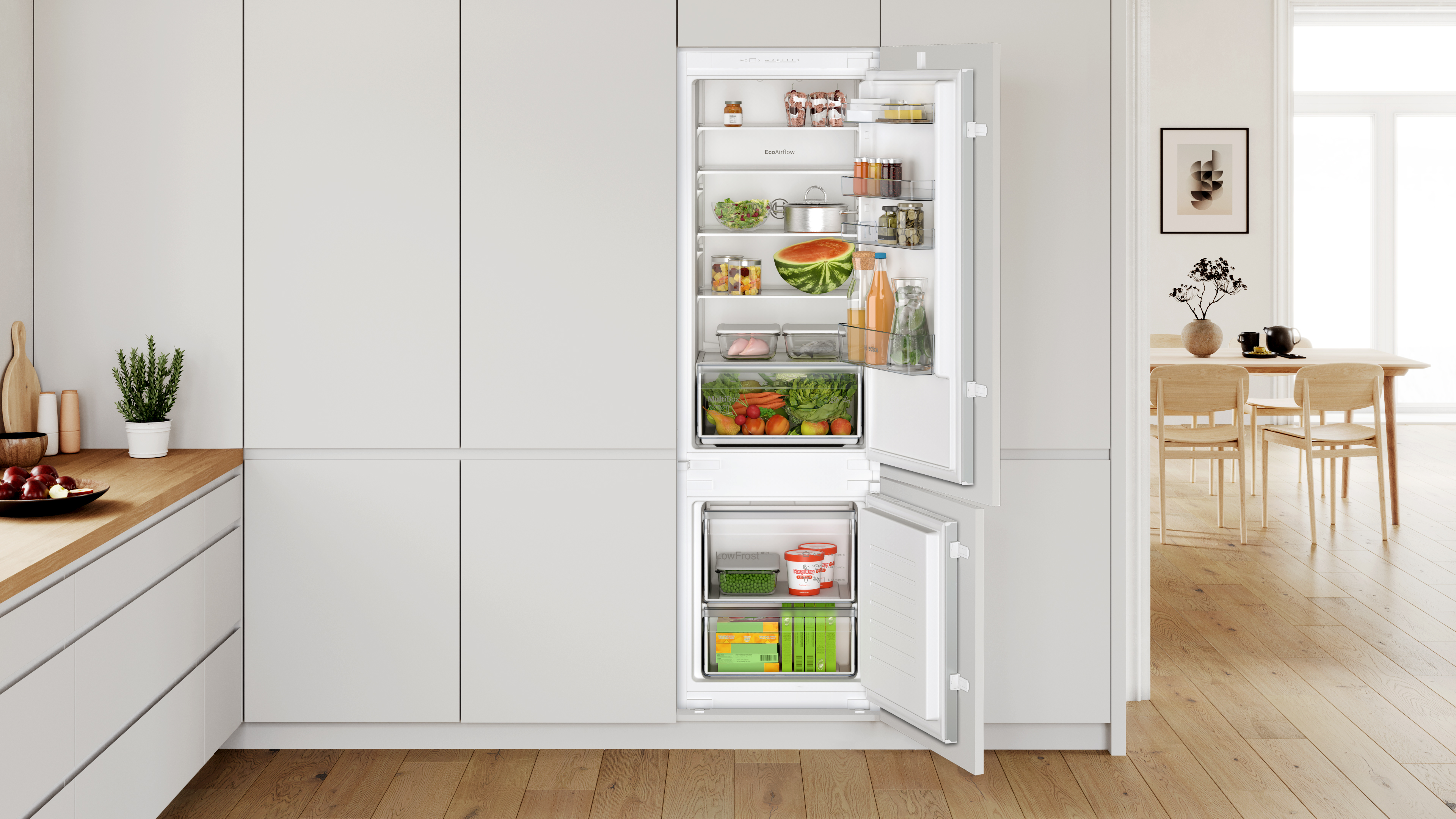 Open refrigerator in a modern gray kitchen shelves show watermelon half salad jars bottles and fresh produce beside a dining area Globalmtc