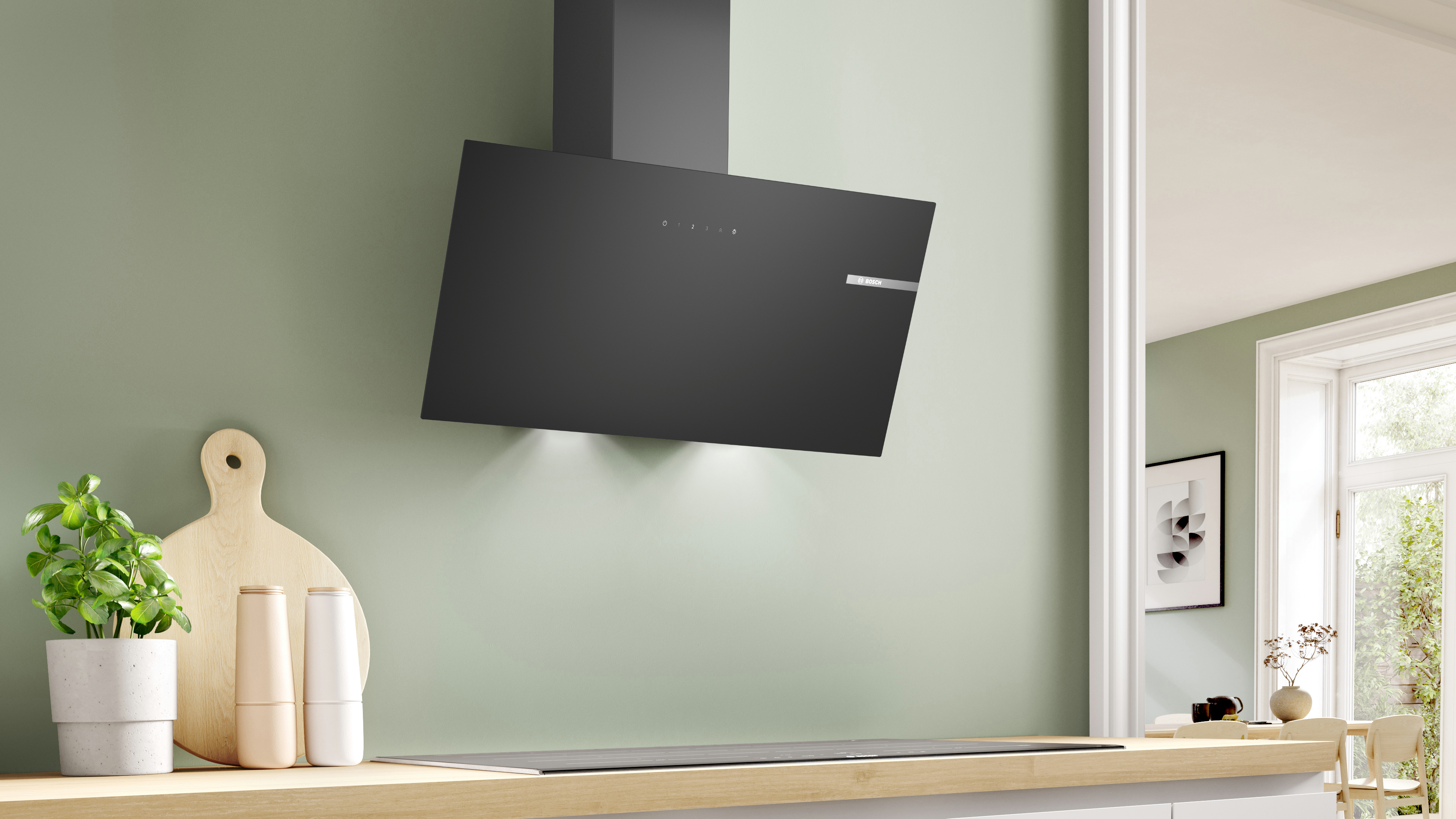 Wall-mounted angled black range hood above a light wood counter in a green kitchen, with plant and cutting board nearby.